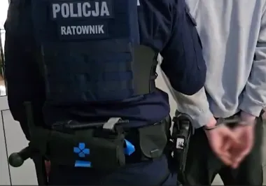Funkcjonariusz policji oznaczony jako ratownik prowadzi zatrzymanego w kajdankach mężczyznę.