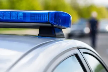 Fragment policyjnego radiowozu z niebieskim światłem na dachu, w tle rozmyta postać człowieka i pojazdy na drodze.